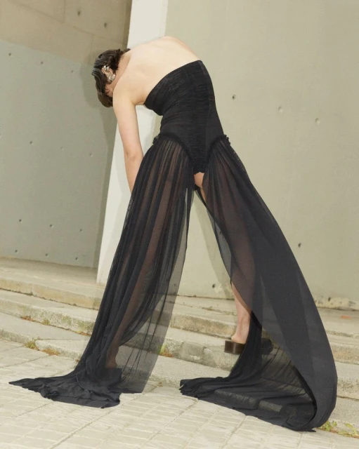 Fashion shoot of a slender adult woman in a flowing strapless black dress against a minimalist concrete outdoor setting.