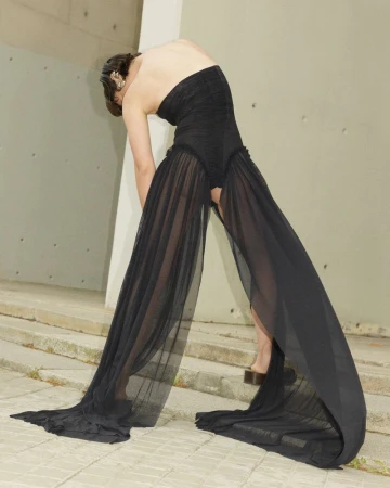 Fashion shoot of a slender adult woman in a flowing strapless black dress against a minimalist concrete outdoor setting.