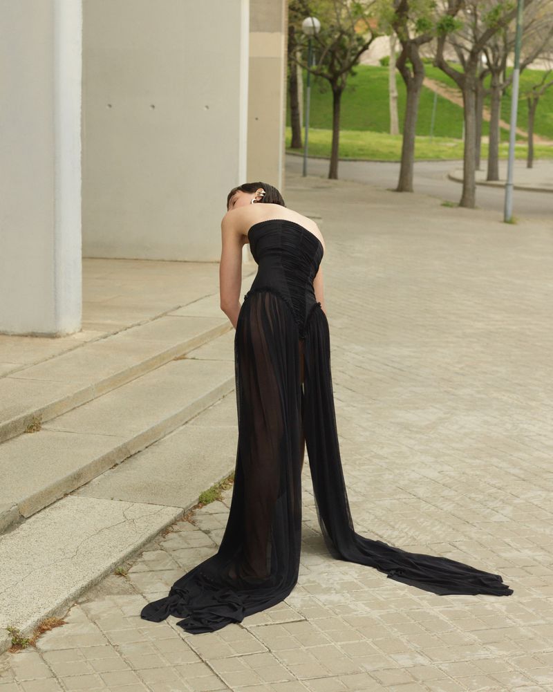 Model in a black strapless gown with long sheer train standing on paved street outdoors.