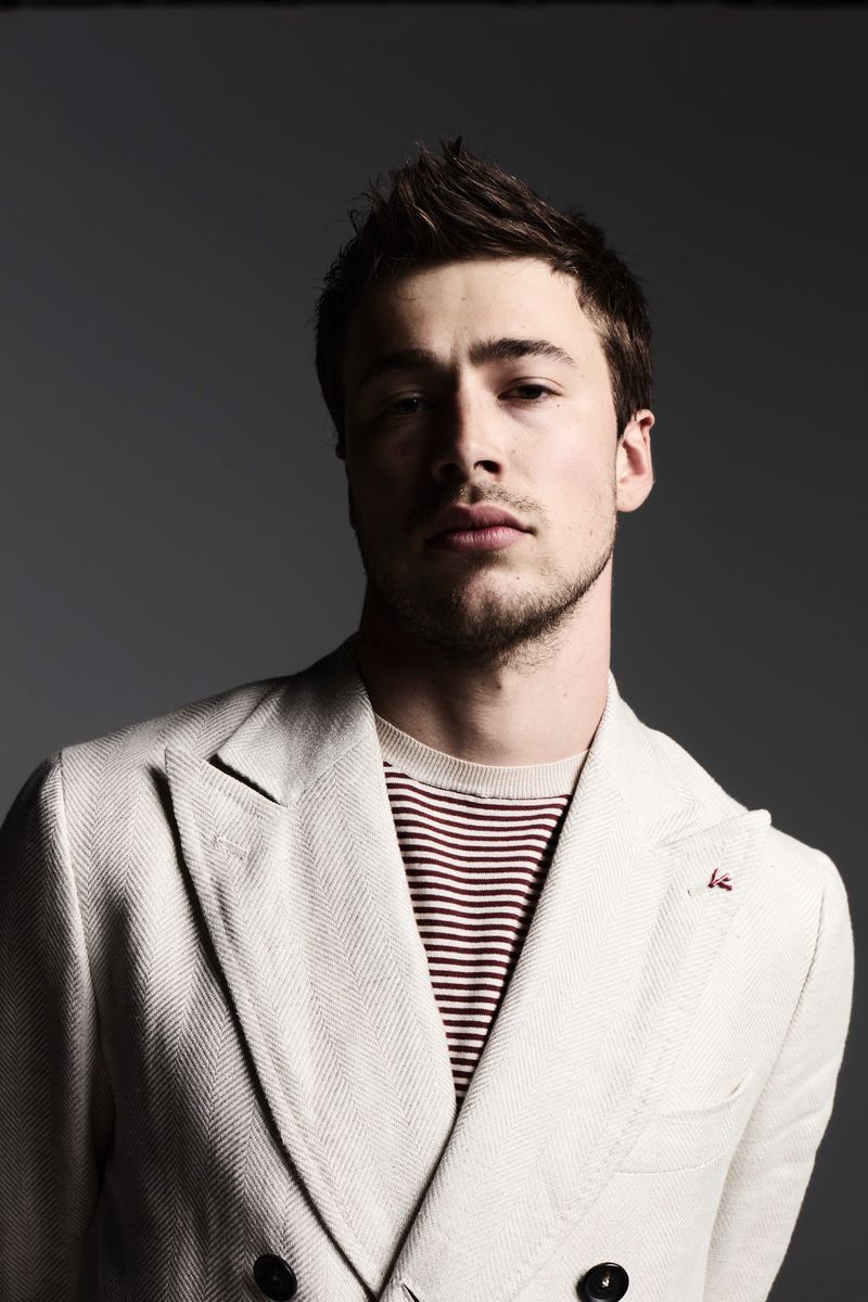Headshot of a young adult male with spiky dark hair and a light beard, wearing a white blazer over a striped shirt in a studio.