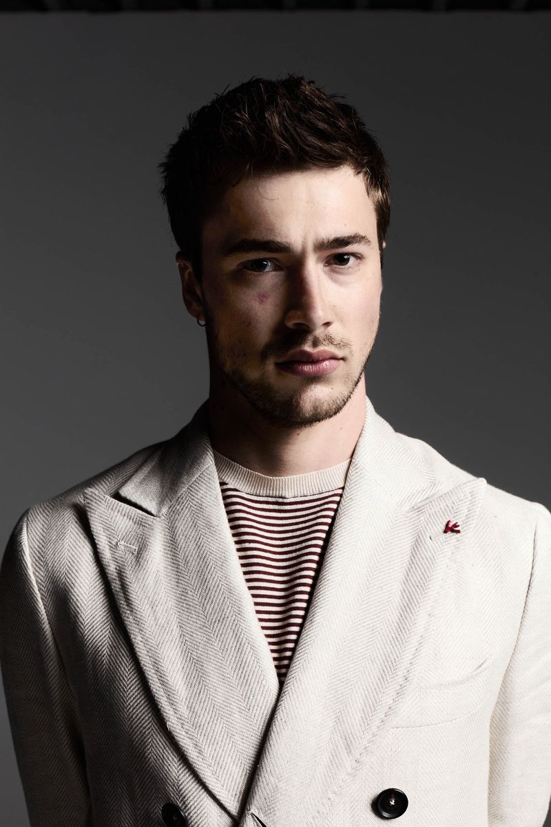 Portrait of a young man in a cream-colored blazer and striped shirt against a grey backdrop, emphasizing fashion and a neutral color palette.