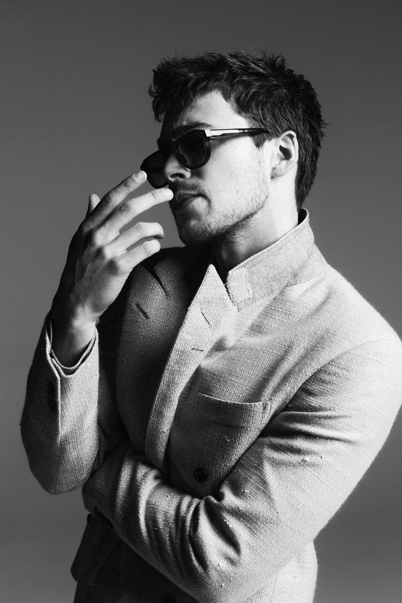 Black and white portrait of a stylish adult man with sunglasses wearing a textured blazer jacket in a studio setting.