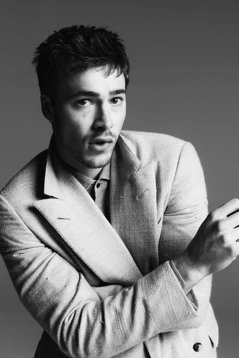 Monochrome portrait of a young adult male in a textured suit, arms crossed, standing against a grey backdrop, looking at the camera.
