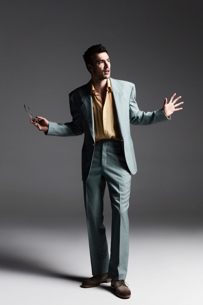 A full-shot studio portrait of a man in a teal suit and glasses, arms out, lit by soft studio lighting against a dark gray background.