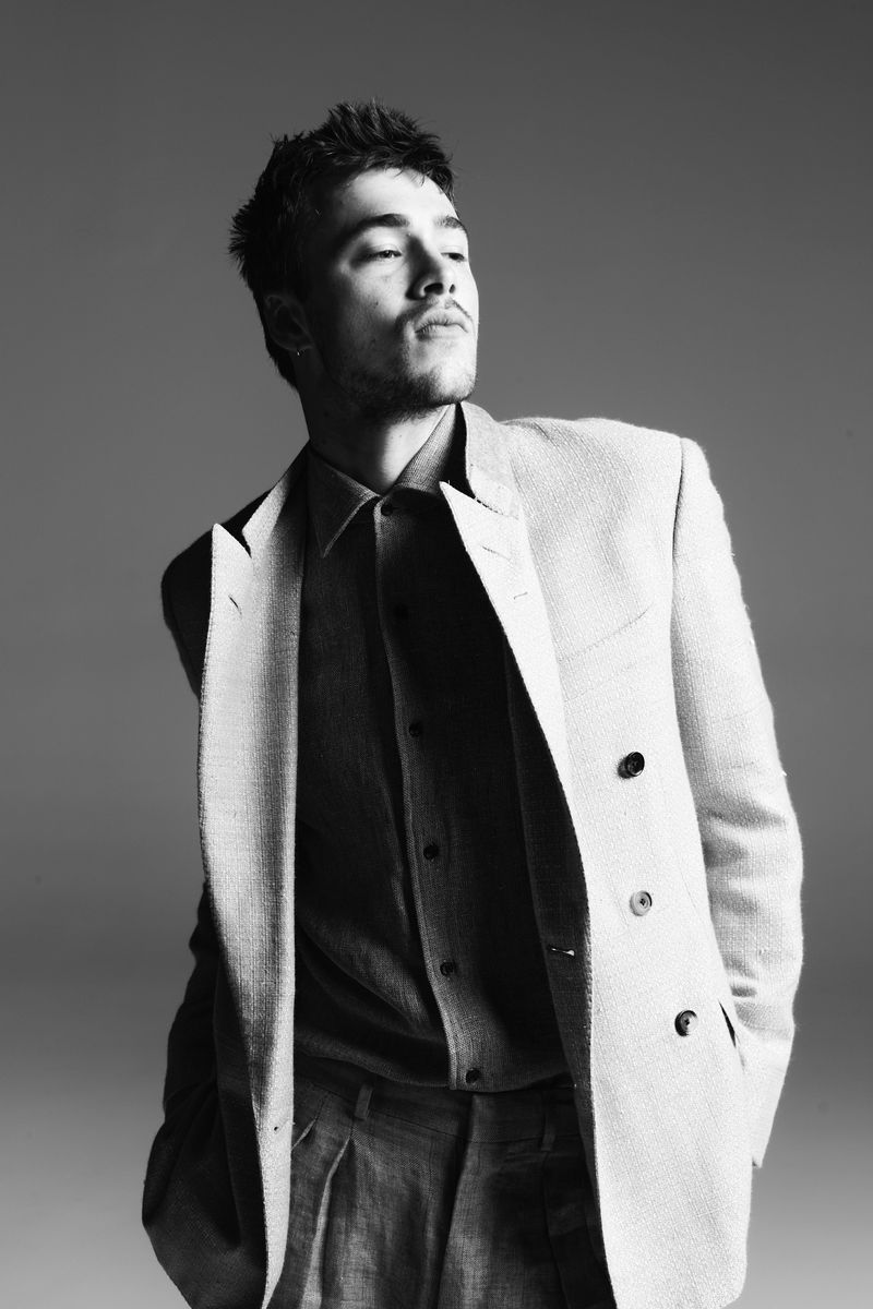 Portrait of a young adult man in a blazer and button-down shirt, shot in black and white with a solid gray backdrop, looking off-camera.