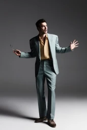 Man wearing a light blue suit and yellow shirt holding glasses against a grey studio backdrop