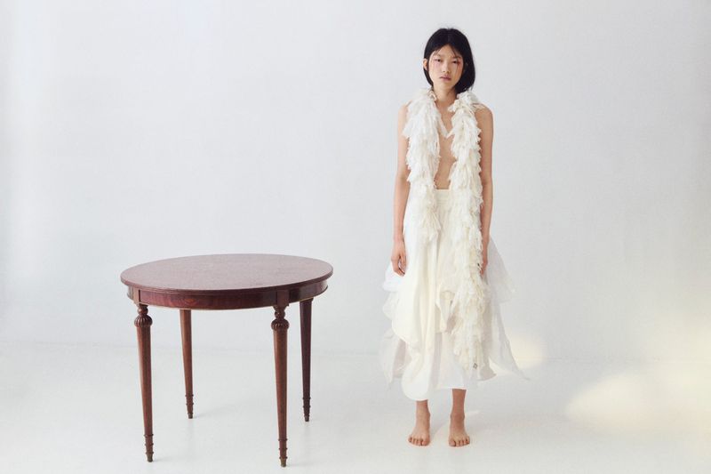 A young Asian woman in a white dress stands barefoot near a round wood table against a white backdrop in a fashion studio shot.