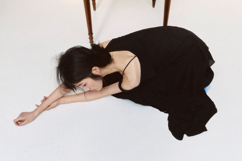 Young woman in black dress poses gracefully on a white floor in a serene indoor setting, casting a minimalist and fashion-forward aesthetic.