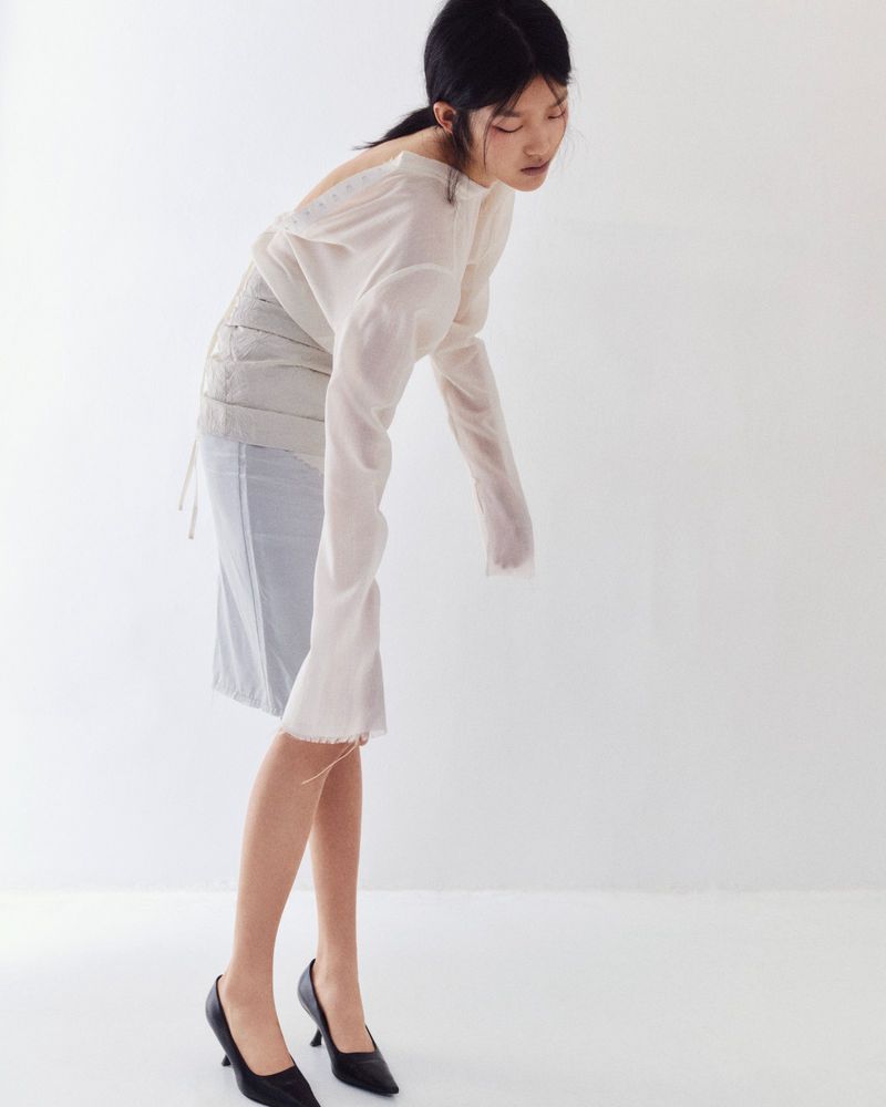 Asian woman in a light-colored, loose-fitting blouse and a light blue pencil skirt, posing in black heels in a bright studio.