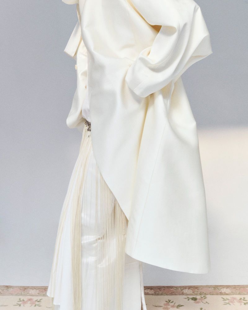 Elegant full-length fashion shot of a woman wearing a satin jacket, fringe skirt, and statement earrings in a light-filled neutral setting.