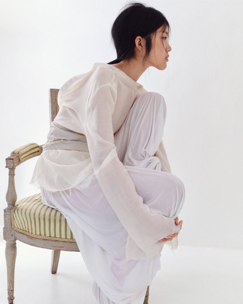 Young woman with dark hair in white sheer top and pants sitting on antique chair in white studio setting looking off to the side.