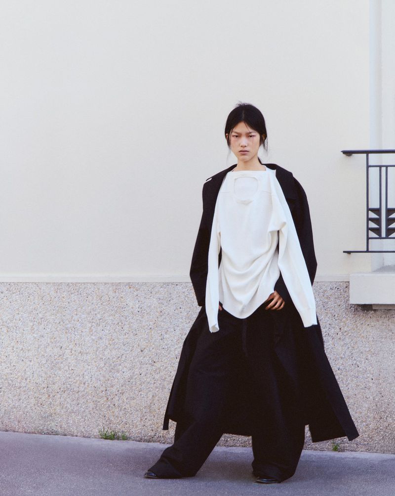 Asian woman in long black coat and white top poses outdoors. Fashion editorial shot against a plain concrete wall backdrop.
