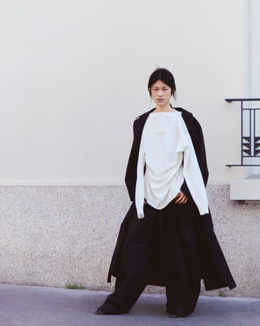 Asian woman in long black coat and white top poses outdoors. Fashion editorial shot against a plain concrete wall backdrop.