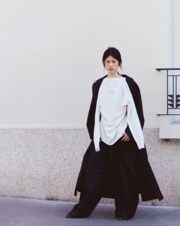 Asian woman in long black coat and white top poses outdoors. Fashion editorial shot against a plain concrete wall backdrop.