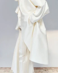 Model wearing a white coat and matching trousers against a plain grey studio backdrop