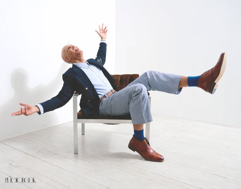 A bald man in a blue blazer, light blue shirt, and striped pants reclines in a chair with arms raised in a white indoor studio setting.
