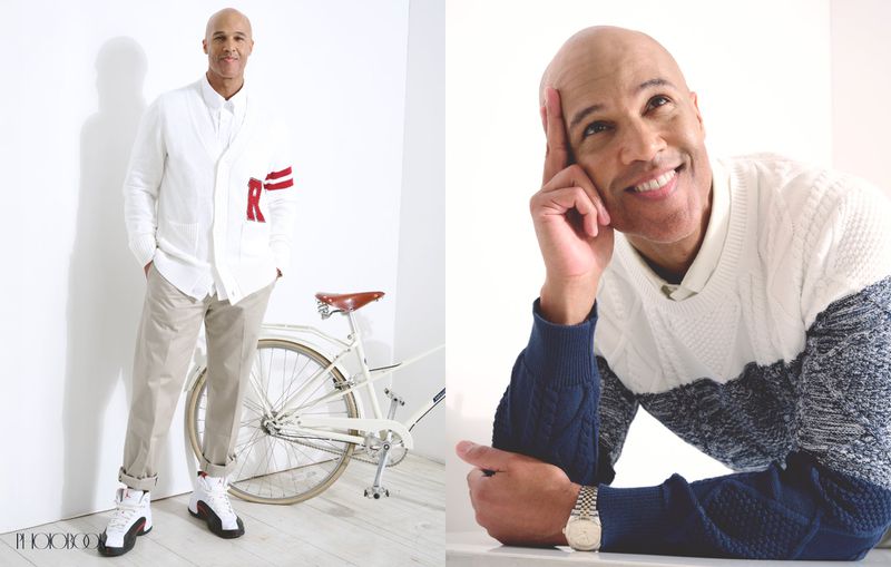 Bald adult man poses in a photo studio with a vintage bicycle, wearing a white cardigan, button-down shirt, khakis, and Air Jordan sneakers.