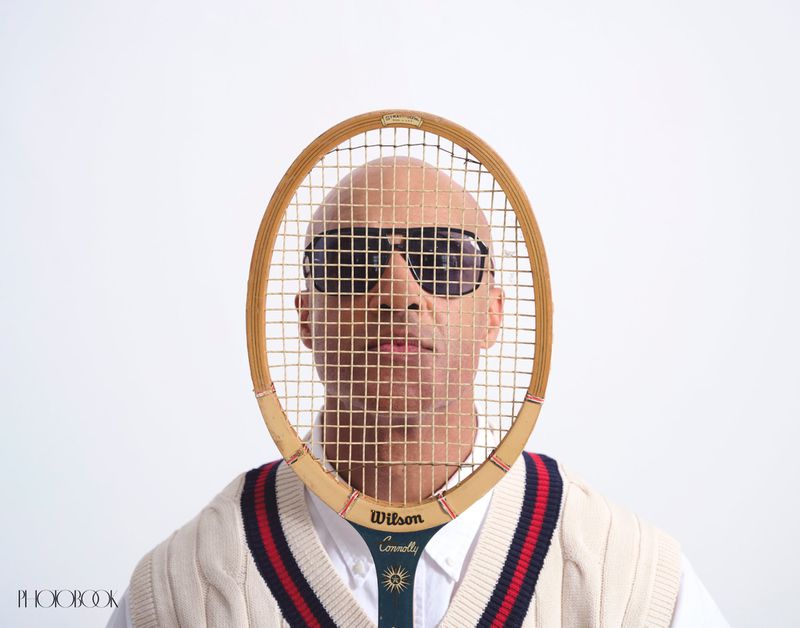 A bald man with sunglasses is framed by a Wilson tennis racket against a plain white background, wearing a cable knit tennis sweater.