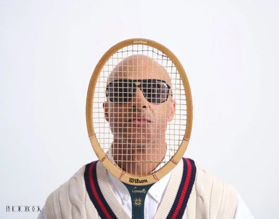 A bald man with sunglasses is framed by a Wilson tennis racket against a plain white background, wearing a cable knit tennis sweater.
