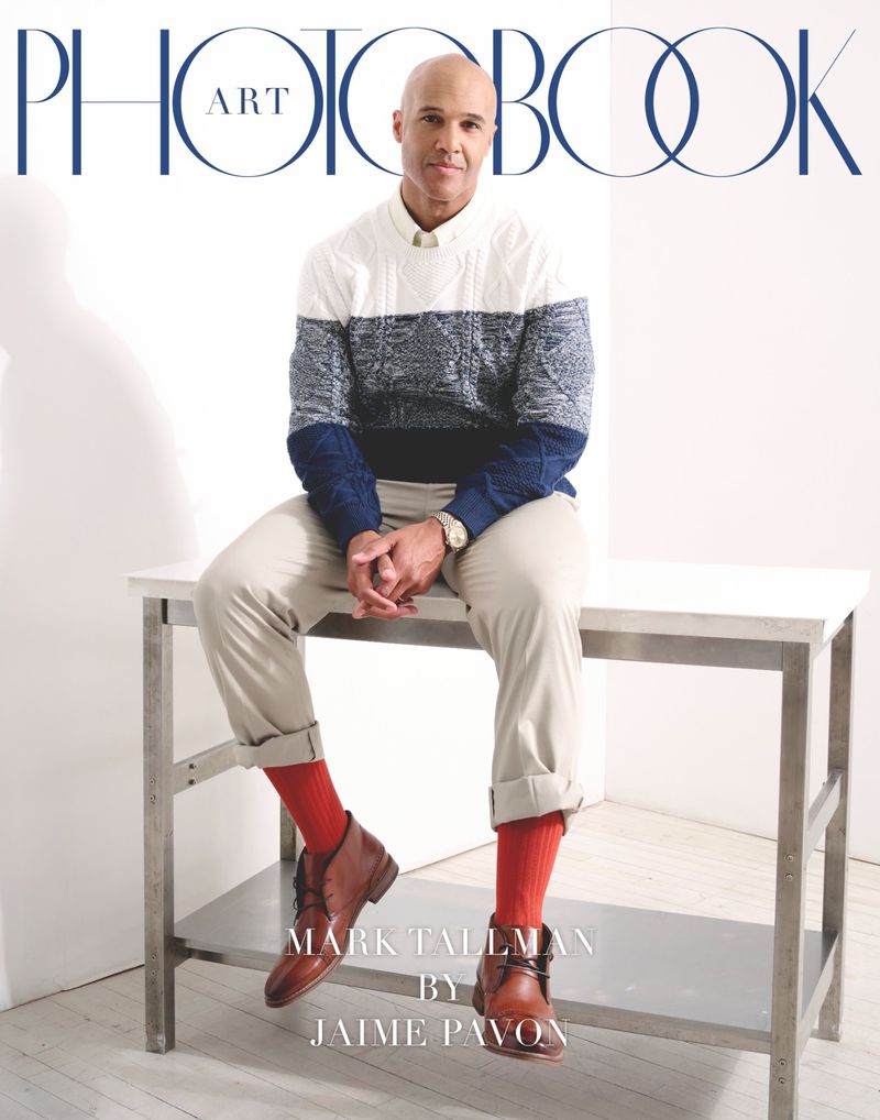 Portrait of a bald man sitting on a metal table wearing a sweater, khaki pants, brown leather boots, and red socks.  Photobook cover.