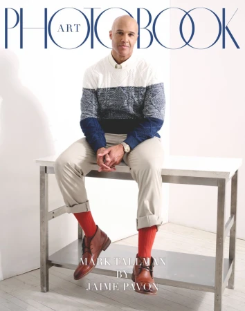 Portrait of a bald man sitting on a metal table wearing a sweater, khaki pants, brown leather boots, and red socks.  Photobook cover.