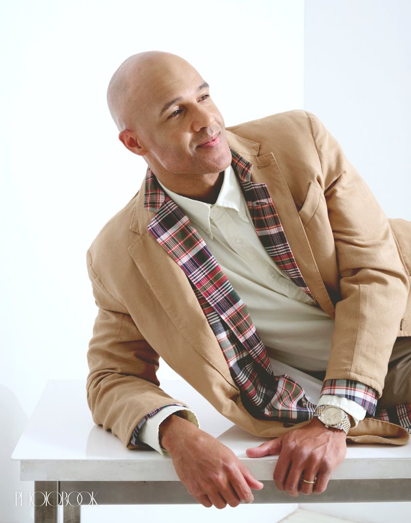 Bald adult male wearing a tan jacket with plaid lining sits on a white table in a brightly lit photo studio, looking off to the side.
