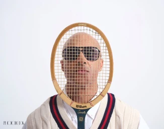 Man wearing sunglasses and a knit vest holding a vintage tennis racket framing his face against a white background.