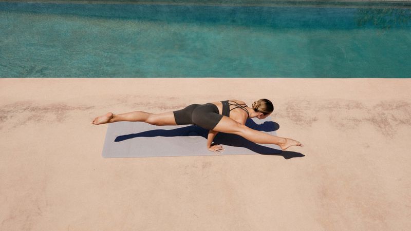 Adult woman in black athletic wear balances in a yoga pose on a grey mat next to a turquoise pool on a tan patio in sunlight.