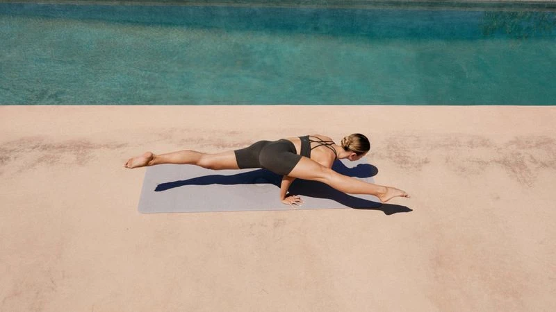 Adult woman in black athletic wear balances in a yoga pose on a grey mat next to a turquoise pool on a tan patio in sunlight.