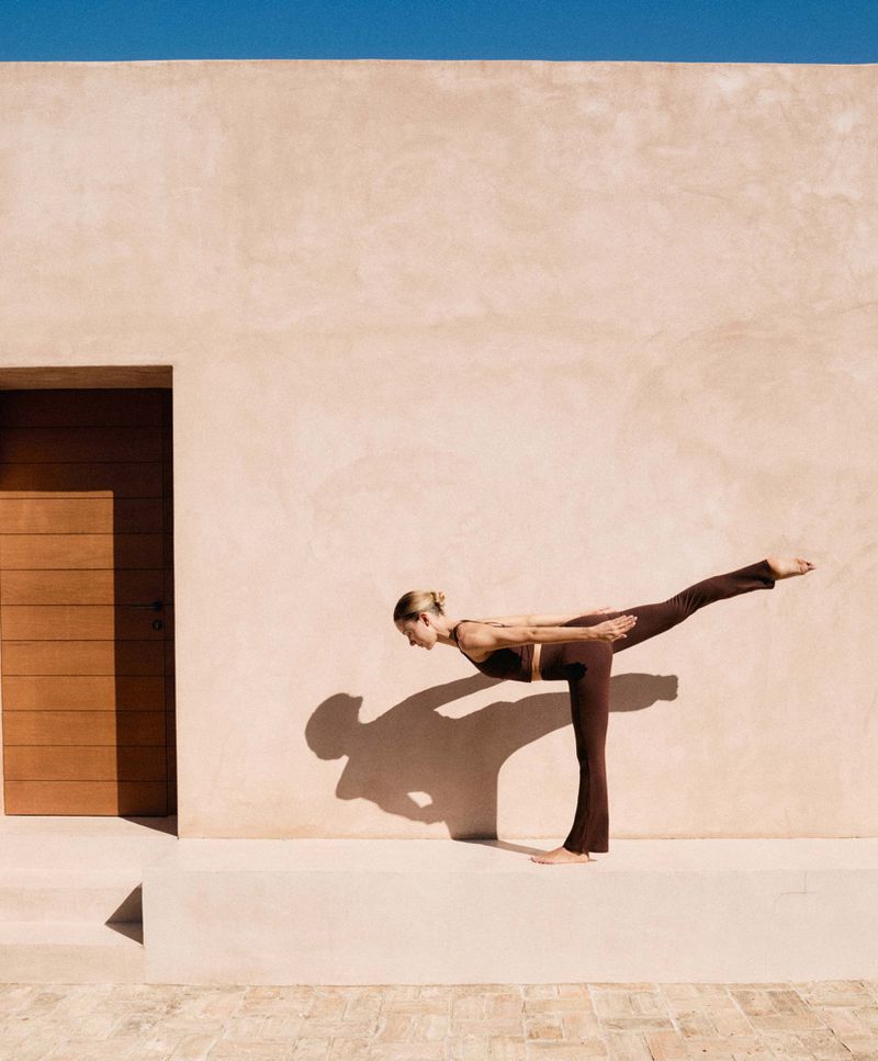 Woman in brown workout gear performs a yoga pose outdoors against a light-colored wall under a clear blue sky in natural sunlight.