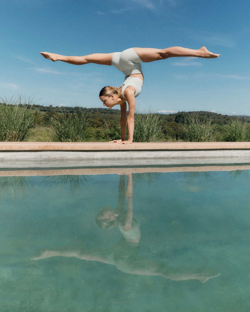 Woman in athletic wear balances in a handstand near the edge of a pool, reflecting in the water against a blue sky and grassy background.
