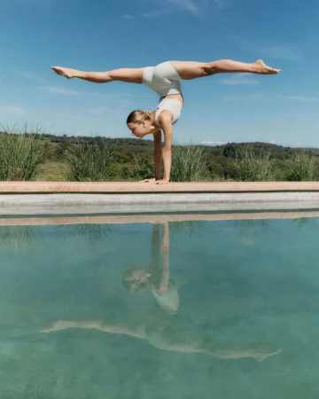 Woman in athletic wear balances in a handstand near the edge of a pool, reflecting in the water against a blue sky and grassy background.