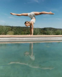 Woman in activewear performing a handstand split by a pool with her reflection visible in the water under a clear sky.