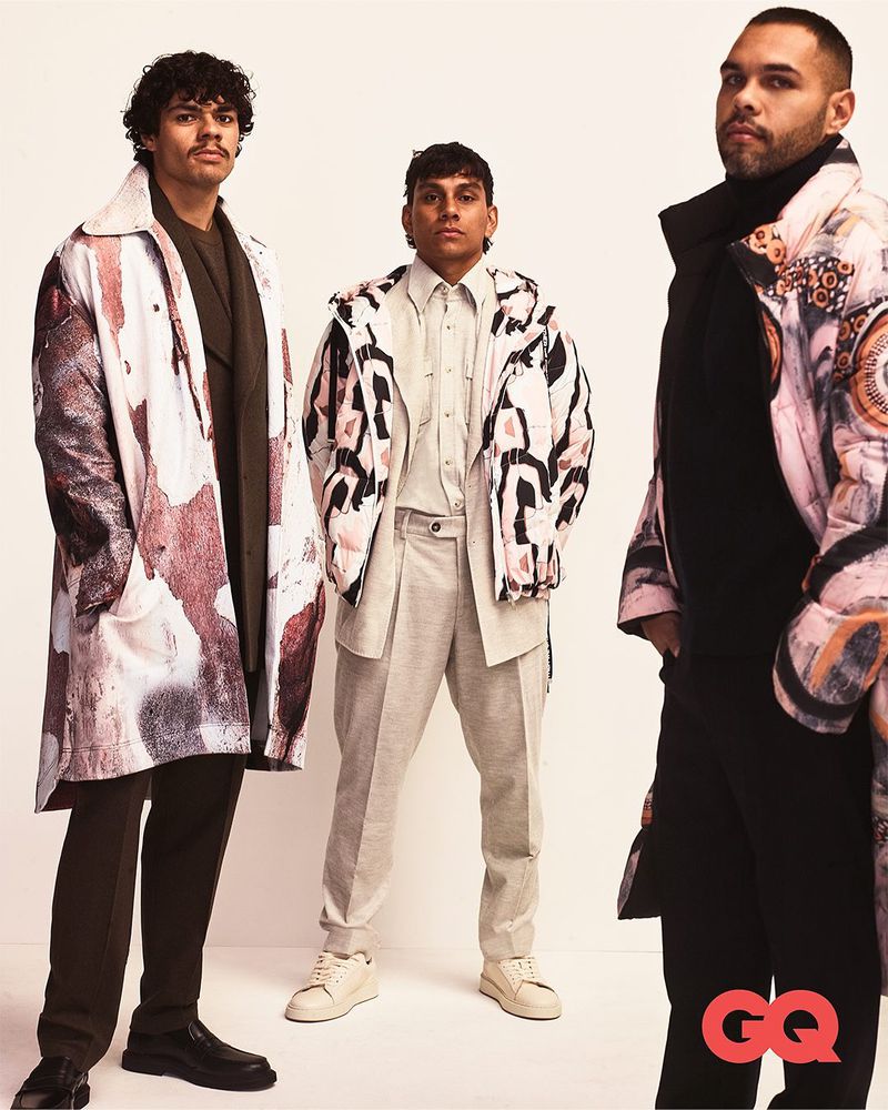 Three male fashion models pose indoors against a neutral backdrop, showcasing patterned outerwear in a studio shot for a GQ editorial spread.