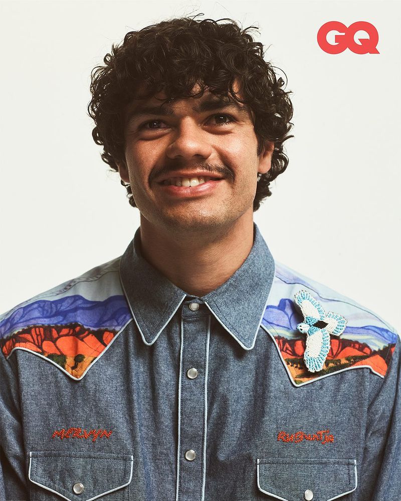 Portrait of a smiling adult male with curly hair and a mustache, wearing a denim shirt with desert landscape details and a bird pin.