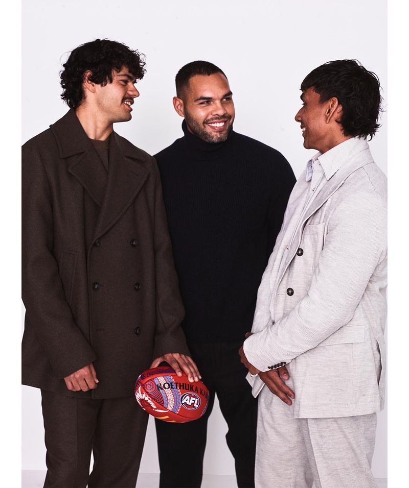 Three adult males in a photo studio wearing fashionable jackets and sweaters posing with an Australian Rules Football (AFL) football.