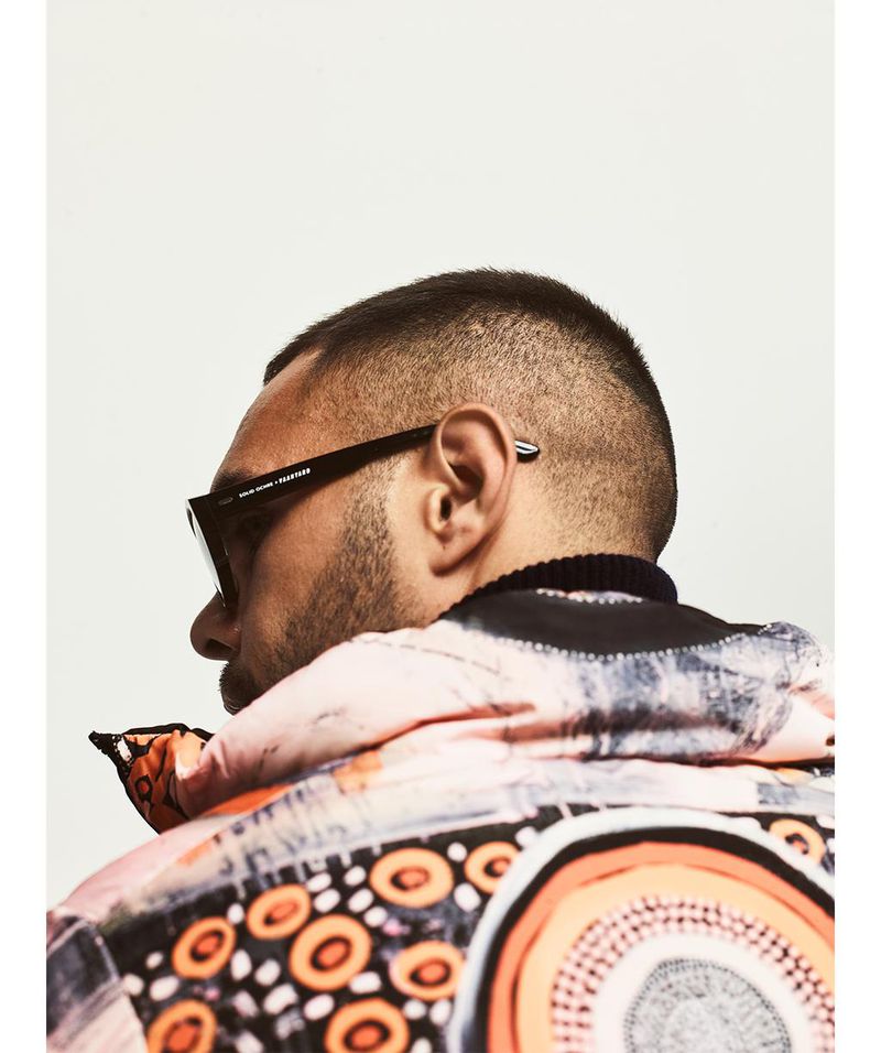 Portrait of an adult male wearing sunglasses and a patterned jacket against a white background in a brightly lit studio setting.