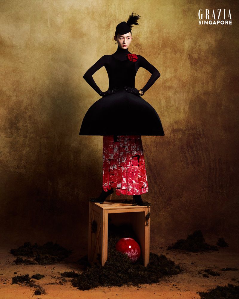 Fashion editorial shot in a studio featuring a woman in a black top, avant-garde black skirt, and a red patterned skirt, posing on a box.