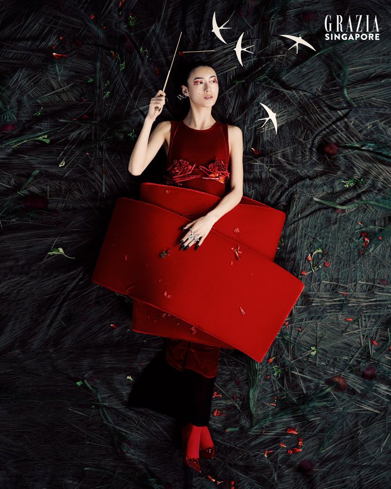 Fashion editorial image featuring a slender Asian woman in a red dress, lying on a textured black surface with scattered rose petals.
