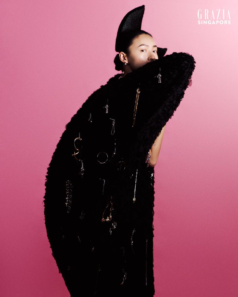 Portrait of a woman with fair skin posing against a bright pink background, wearing a black textured coat with dangling jewelry.