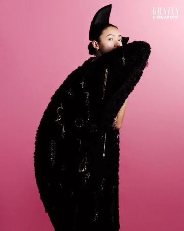 Portrait of a woman with fair skin posing against a bright pink background, wearing a black textured coat with dangling jewelry.