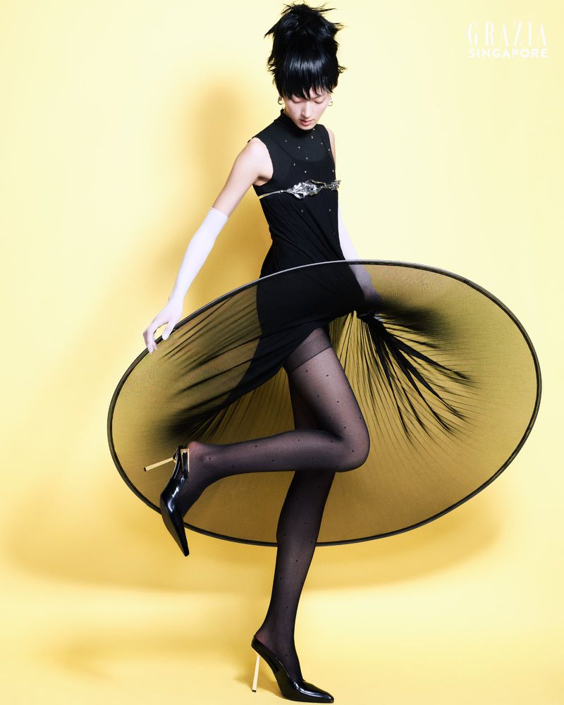 Asian female fashion model in a black dress, gloves, polka-dot stockings, and black heels, posing in front of a yellow backdrop.