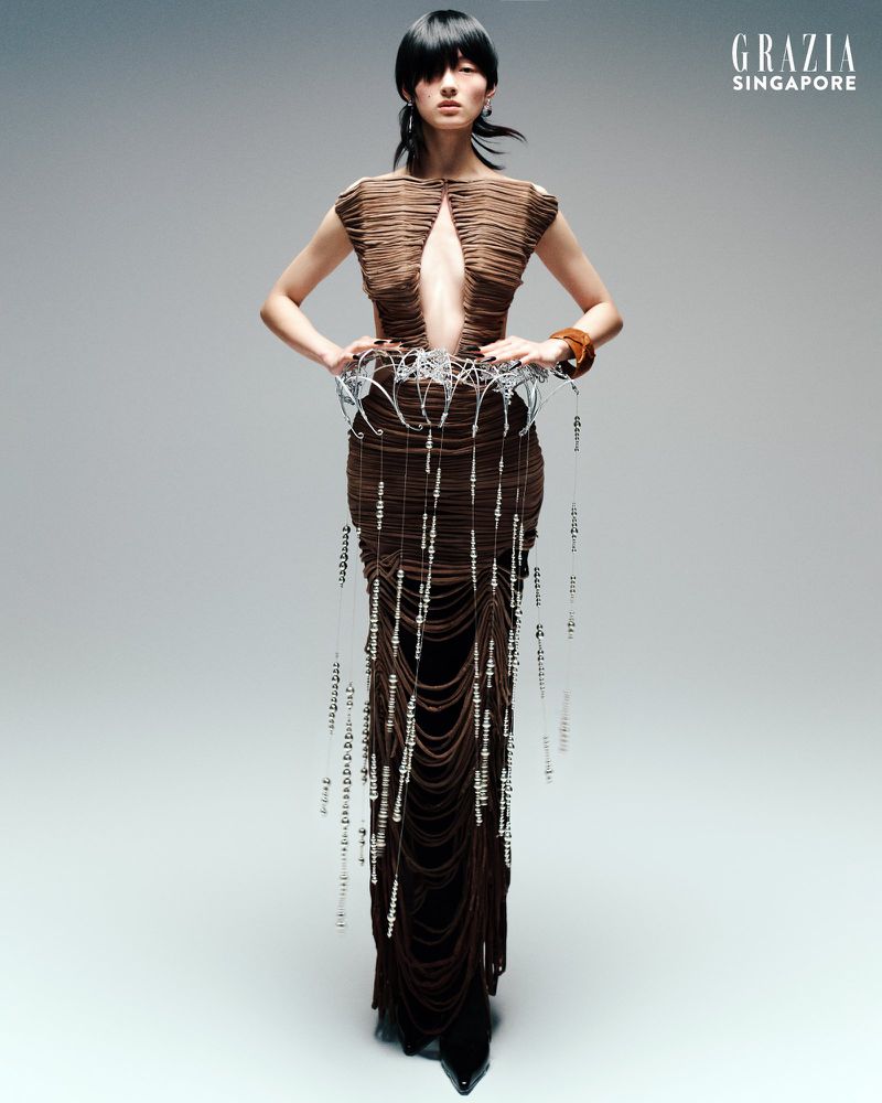 Fashion shot of an Asian woman wearing a sculptural brown dress with beaded details, posing against a gray background for Grazia Singapore.