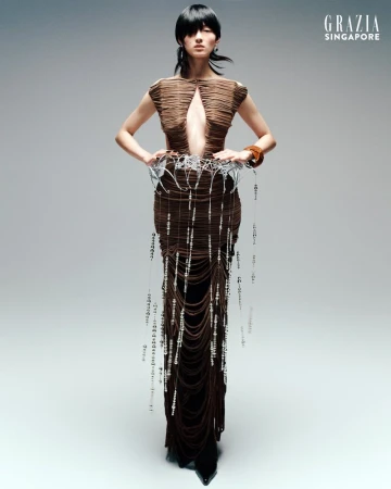 Fashion shot of an Asian woman wearing a sculptural brown dress with beaded details, posing against a gray background for Grazia Singapore.