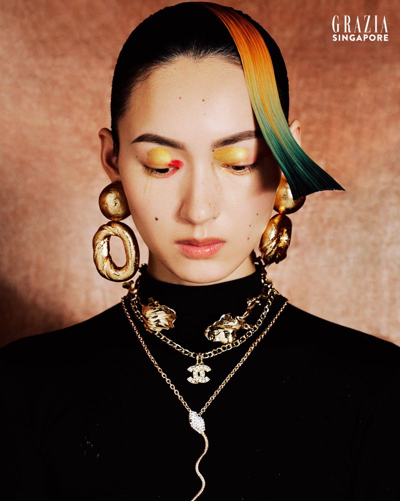 Portrait of a woman with striking hair and makeup, adorned with gold jewelry, against a textured brown backdrop; fashion-forward beauty.