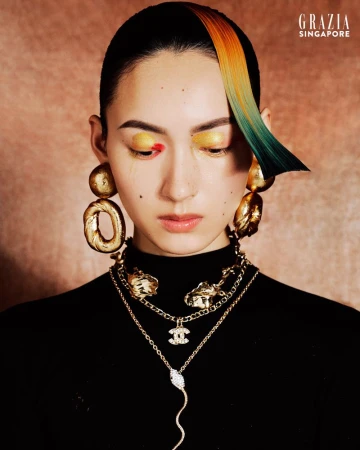 Portrait of a woman with striking hair and makeup, adorned with gold jewelry, against a textured brown backdrop; fashion-forward beauty.