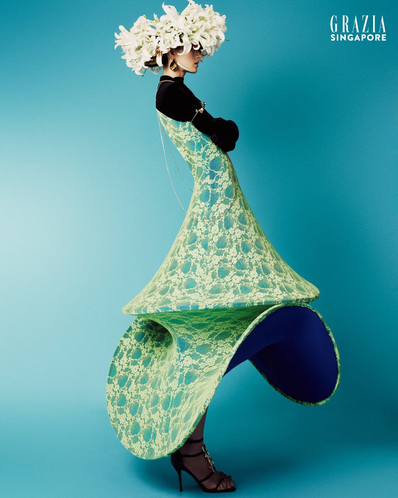 Fashion editorial shot of a woman in a sculptural light green lace dress with flowers on her head against a teal background in studio.