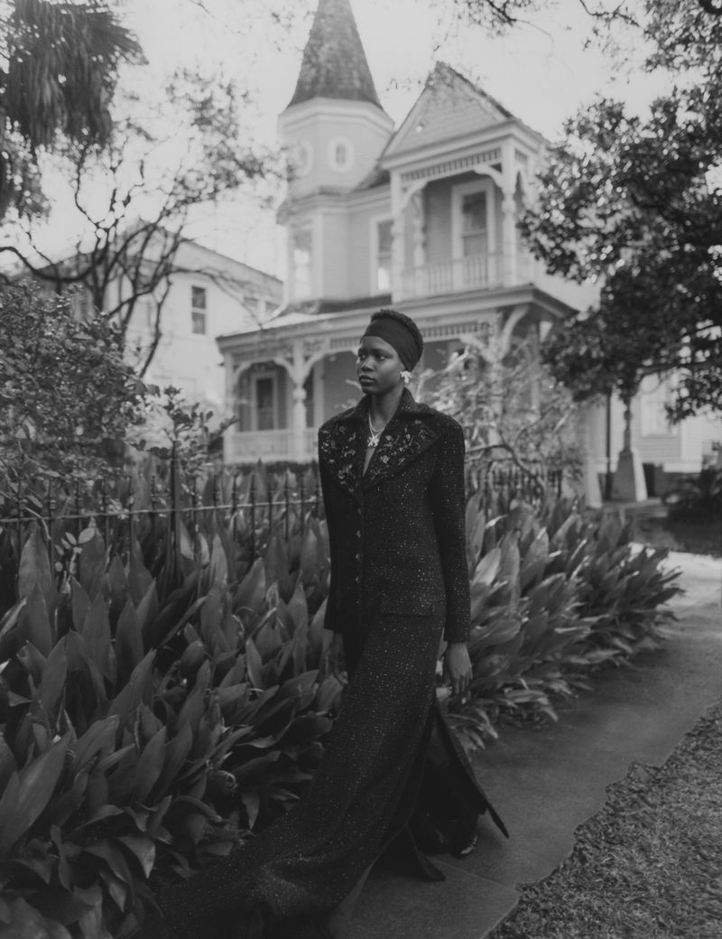 Black and white outdoor shot of a Black woman in a dark, stylish suit walking in front of a house with lush green foliage.
