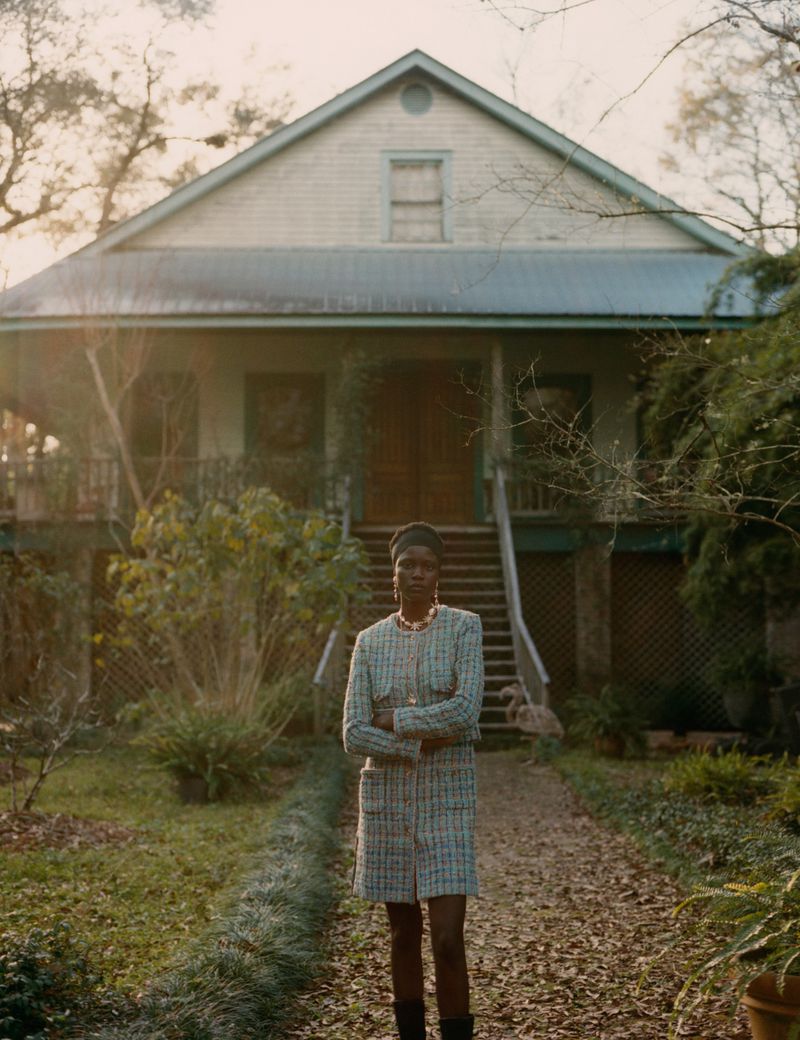 A fashionable woman in a tweed skirt suit stands in front of a rural house with a long, tree-lined driveway and manicured lawn.