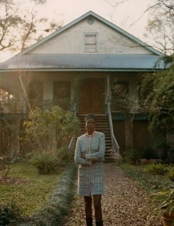 A fashionable woman in a tweed skirt suit stands in front of a rural house with a long, tree-lined driveway and manicured lawn.
